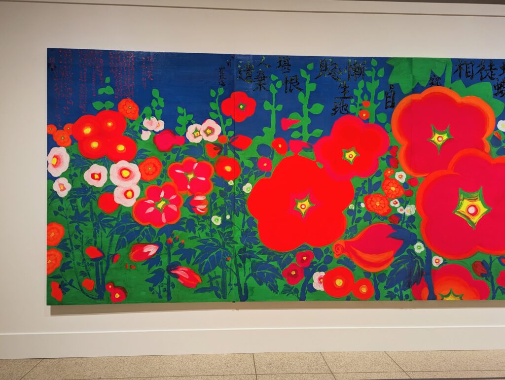 Photo of the left 60% ish of the painting "Standing on the Hollyhock Field" by Hong Jiyoon. It's a very large, horizontal painting of big red, pink, and white flowers on a green and blue background. There is red and black hanja writing at the top. The colors are very vivid. A little bit of white wall and beige tile floor are visible at the edges of the photo.